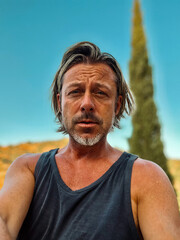 Tanned man with blond half long hair and stubble beard in singlet shirt in sunny countryside with...