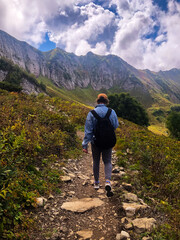 Fototapeta premium Man with a backpack hiking along the Path of Health trail at Rosa Khutor resort in Sochi, Russia, seen from behind. Concept of outdoor adventure, active travel, and exploring alpine landscapes