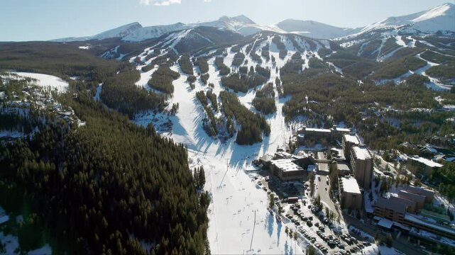 An aerial view of the popular Breckenridge ski resort in Colorado, with snow covered mountain peaks and high altitude trails full of winter skiers and snowboarders on a sunny winter day.