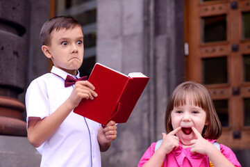 Two children having fun in front of school doors. Boy with jokingly serious face reading from...