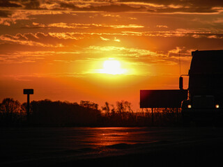 A vibrant sunset casts a warm glow over the highway, silhouetting a large truck against the fiery...
