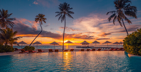 Stunning sunset view of infinity pool with canopy chairs, loungers, and umbrella under palm trees facing sea tropical resort setting for summer vacation, leisure, relaxation beautiful travel landscape