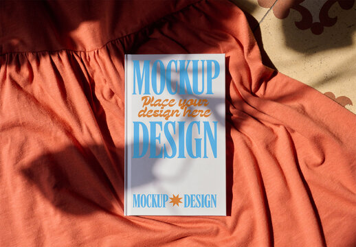 A blank notebook cover rests on soft fabric. Shadows from surrounding objects create interesting patterns on the surface. PSD template mocky&iexcl;up