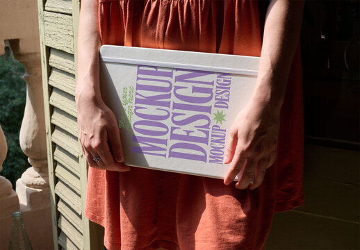 Anonymous Woman holds a minimalist notebook cover in their hands, standing on a balcony surrounded by greenery in bright sunlight. PSD template mockup