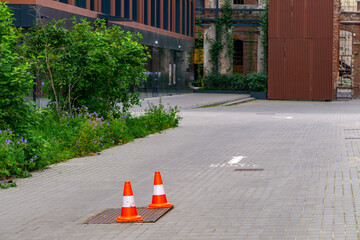 Two orange traffic cones placed on a paved surface near a utility hatch. Modern urban surroundings...