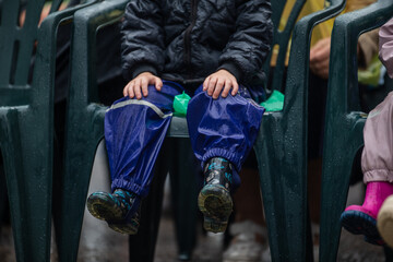 children sitting on plastic chairs during outdoor event in rainy weather wearing waterproof pants and rubber boots, close-up of kids legs in colorful rain gear on wet day at festival or performance