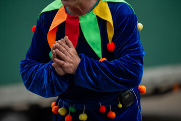 clown performer in colorful velvet costume with pom-poms holding hands together, festive jester...