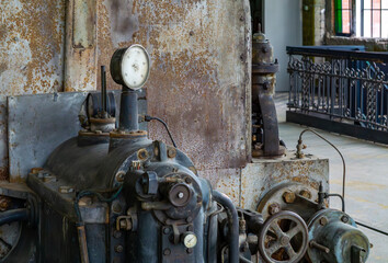 Vintage industrial machine with a pressure gauge, valves, and pipes, located in an old factory hall with a metal railing and colorful windows. Visible signs of wear and rust add to the retro industria