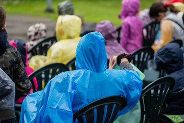 colorful crowd of people in raincoats sitting on plastic chairs during outdoor event rainy day...