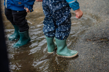 children in green rain boots splashing in water puddle during rainy day on wet asphalt playful...