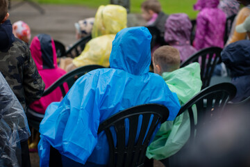 colorful crowd of people in raincoats sitting on plastic chairs during outdoor event rainy day...