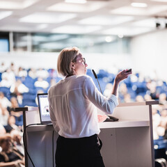 Female speaker giving a talk on corporate business conference. Unrecognizable people in audience at...