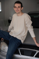 Enthusiastic young man poses casually on silver car in modern garage setting showcasing relaxed...