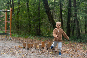 Little boy in brown fluffy jacket jumping on wooden stepping platforms at outdoor forest playground...