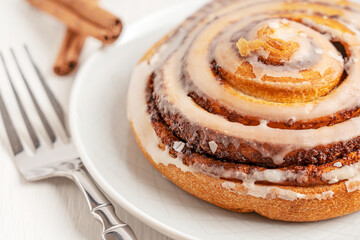 Close-up view of a delicious homemade sweet cinnamon roll bun dessert glazed with sugar icing on a gray plate on white wooden table with silver fork served for morning breakfast as baked pastry snack