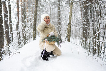 A beautiful woman with short blonde hair is dressed in a white fur coat and black boots. She stands against the background of a snowy landscape with a basket and a bouquet of fir branches.
