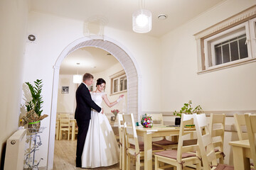 A bride and groom are standing in front of a long dining table with chairs