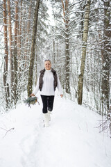 Obraz premium A beautiful woman with blonde hair is dressed in a fur coat and black boots. She stands against the background of a snowy landscape with a basket and a bouquet of fir branches.
