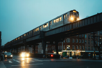 Subway train traveling on elevated line in Brooklyn at dawn  Metro gliding over urban street with headlights and glowing storefronts