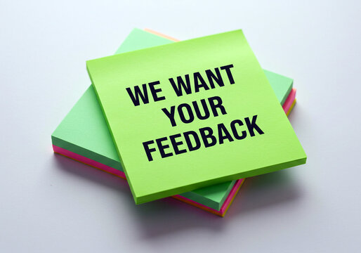 Bright green sticky note with bold black text reading we want your feedback resting on a stack of colorful paper notes against a clean white background