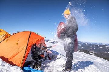 Laughing friends throw snow from inside an orange tent at a smiling man with a sleeping bag on a...