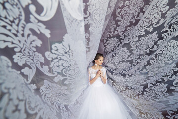 A woman in a white dress is standing in front of a curtain with a lace pattern