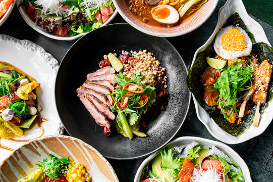 Assorted Asian dishes including ramen, grilled meat, skewers, and salads arranged on a table, showcasing colorful, appetizing food in a restaurant setting.