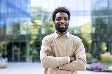 A smiling man with crossed arms stands in front of a modern building. He is dressed casually and...
