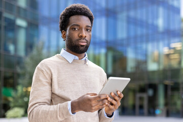 An african american man in a light sweater, holding a tablet, posing in front of an office...