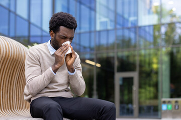 An African-American man blows his nose while seated outdoors in front of a modern building.