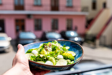 Hand holding a bowl of fresh vegetable salad with broccoli, cucumber, lettuce, and mozzarella balls on a city street background. Outdoor healthy eating concept.