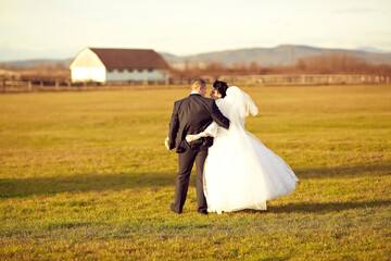 A bride and groom are walking through a park, with the bride wearing a white dress and the groom...