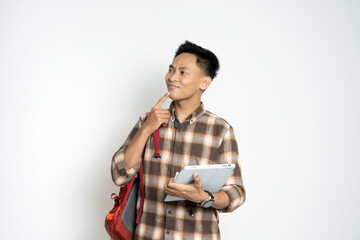 Young student wearing a checkered shirt standing confidently, holding a tablet and a backpack, thinking deeply.