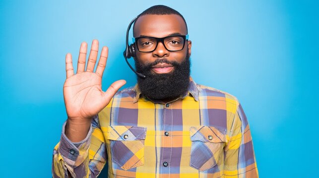 African American man wearing headset and plaid shirt is greeting with raised hand against a blue background, showcasing customer service and communication skills in a professional setting - Powered by Adobe