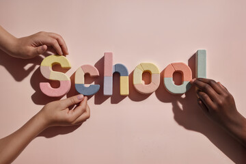 Children's hands arranging the word SCHOOL from colorful geometric shapes against the pastel pink background. Back to school, educational backdrop. 