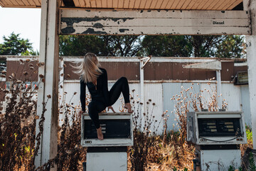 A stylish blonde woman in a black bodysuit poses barefoot on top of an old, weathered gas pump at...