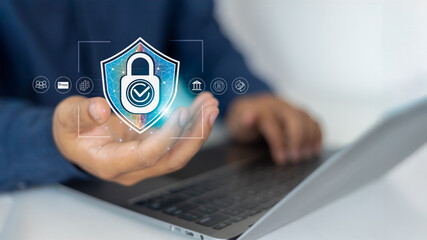 Close-up of a hand holding a digital shield lock icon, with a person typing on a laptop in the...