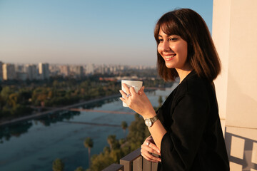 Young woman smiling and drinking some coffee and looking out from balcony
