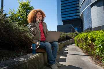 Curly-haired man talking on phone while working on laptop outdoors. Remote work, mobile communication, sustainable lifestyle and modern eco-friendly digital career © LaMorenita