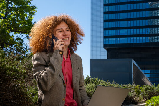 A curly-haired redheaded man smiles while talking on the phone and working on a laptop outdoors. Mobile remote work, positive business communication and sustainable lifestyle