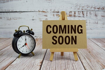 Coming Soon text with easel and alarm clock on wooden background