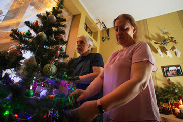 Joyful couple places colorful ornaments on their Christmas tree, surrounded by festive lights in a warmly decorated