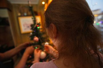 Decorates Christmas tree with colorful ornaments while family shares joyful moments indoors in warm lighting