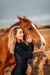 Fototapeta premium Stylish Woman in Black Jumpsuit Standing with Horse near Hay Bales at Sunset