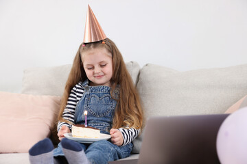 Birthday celebration. Little girl with piece of cake having video call via laptop on sofa at home