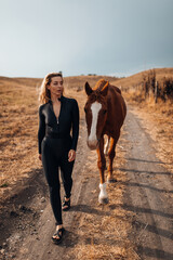 Stylish Woman in Black Jumpsuit Standing with Horse near Hay Bales at Sunset