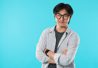 Handsome Asian man in glasses with crossed arms on light blue background. Space for text