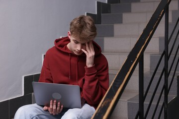 Cyber bullying. Upset teenage boy with laptop on stairs indoors