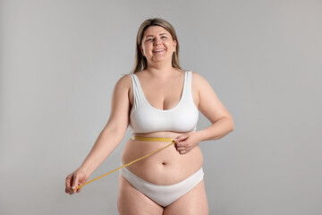 Weight loss. Plus size woman measuring waist with tape on gray background