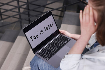 Cyber bullying. Upset teenage girl looking at laptop with text You Loser on stairs indoors, closeup